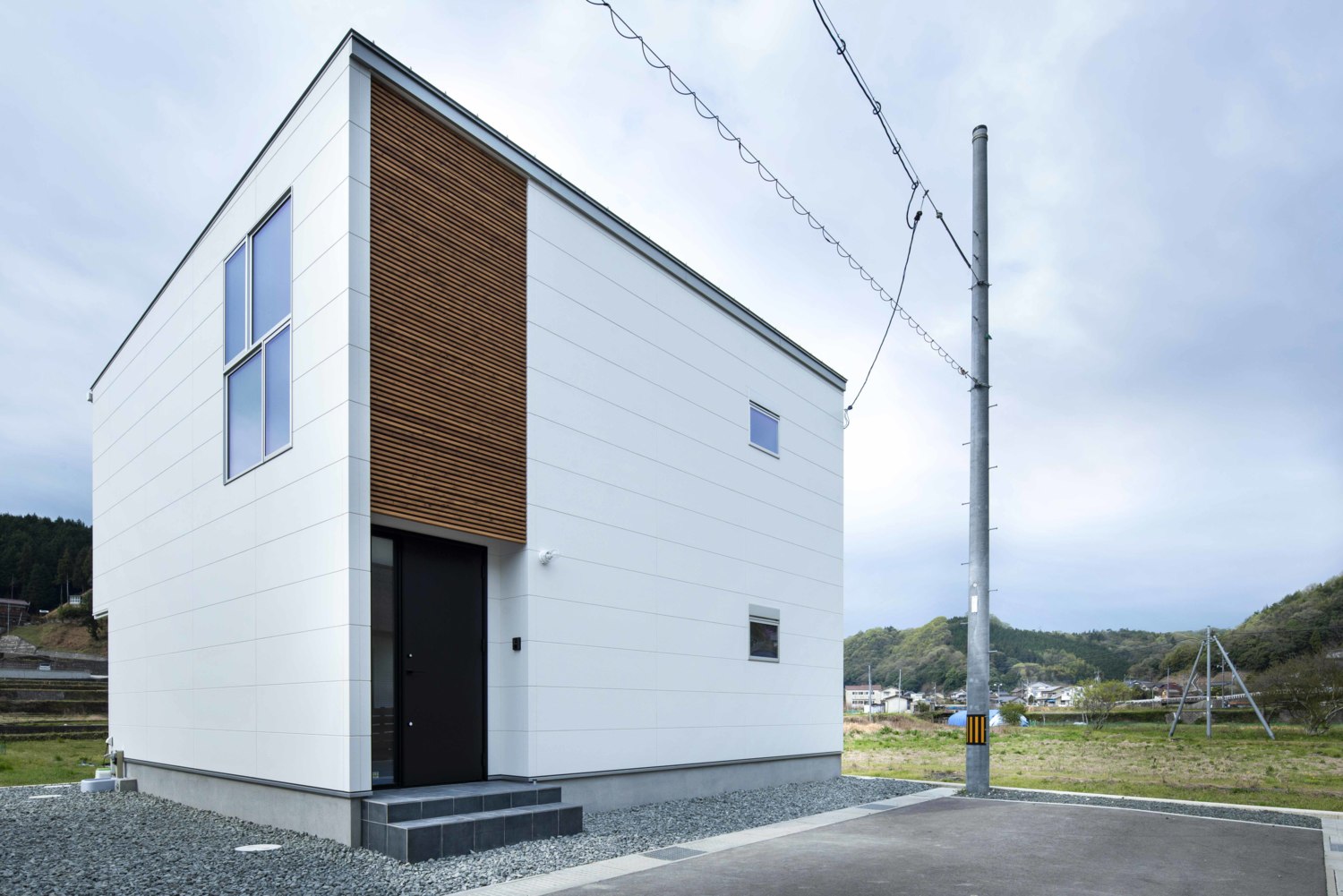 白い箱型のシンプルな形状の建物に木製ルーバーや黒色の玄関ドアが目を引く、若々しく優しいイメージの外観。